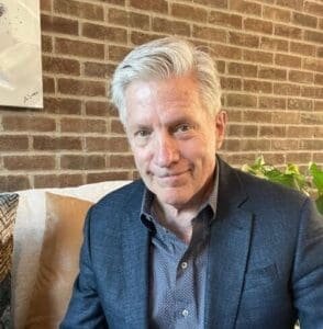A man with gray hair, wearing a dark blazer and patterned shirt, sits on a couch against a brick wall, looking at the camera. Green plants in the background suggest a relaxed environment.