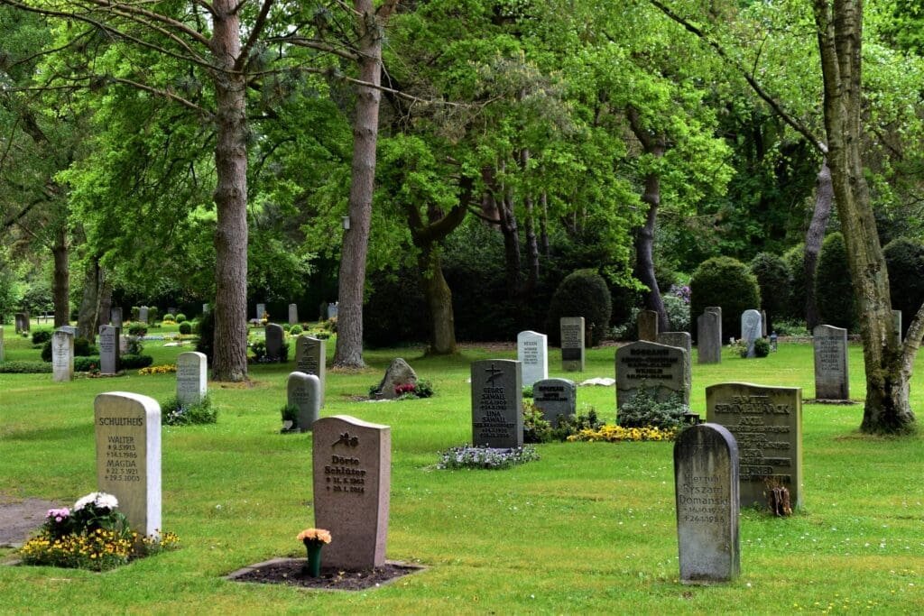 Gravestones scattered across a serene, green cemetery, surrounded by tall trees and flower beds.
