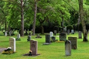 Gravestones scattered across a serene, green cemetery, surrounded by tall trees and flower beds.