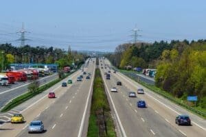 Six-lane highway with flowing traffic, featuring various cars and trucks alongside a service area. Power lines and greenery create a scenic backdrop.