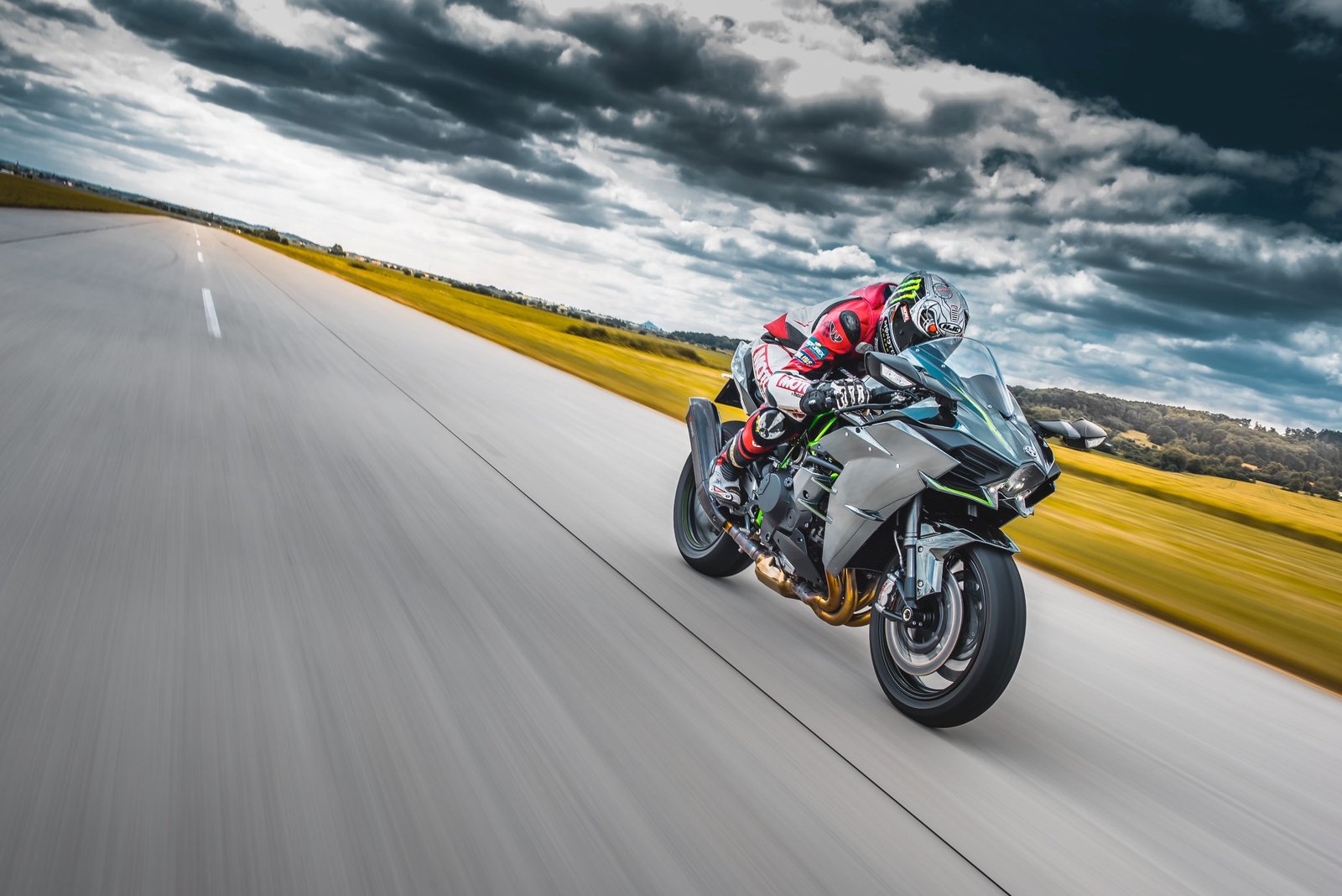 A motorcyclist leans into a curve on a sleek, modern bike, speeding along a two-lane road with an expansive field and dramatic sky behind.