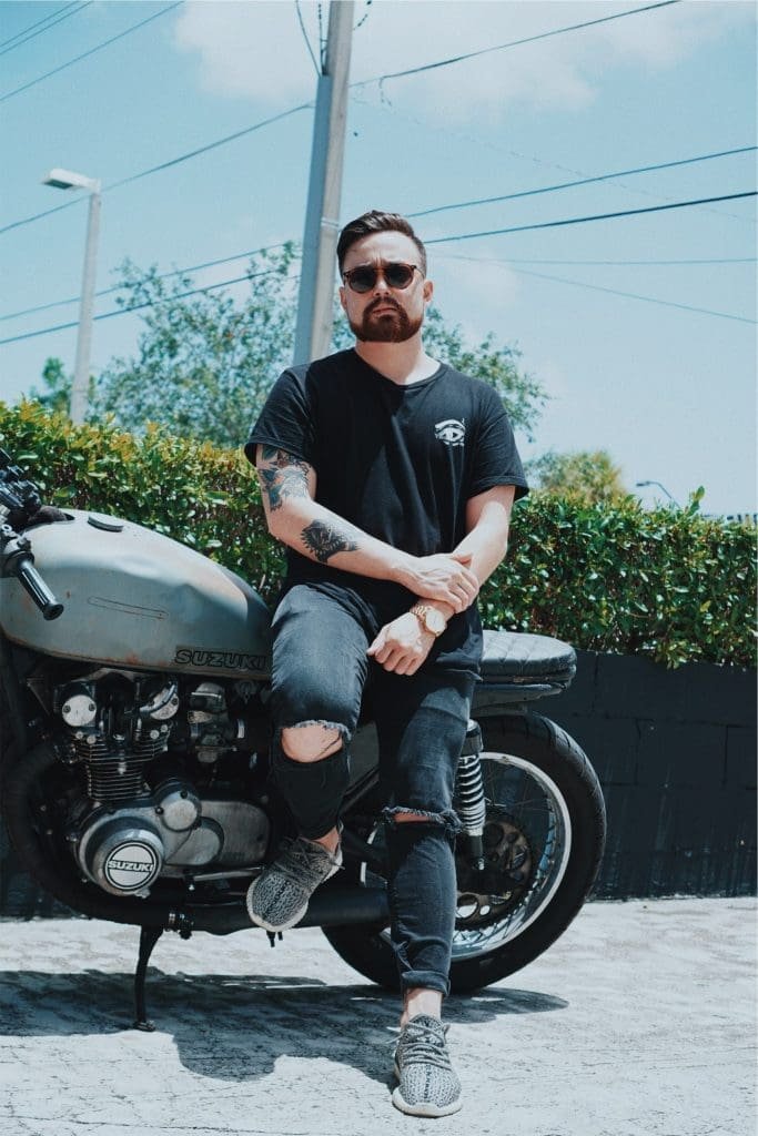 Man with a beard and sunglasses leans casually on a vintage Suzuki motorcycle, wearing a black t-shirt and ripped jeans against a sunny backdrop.