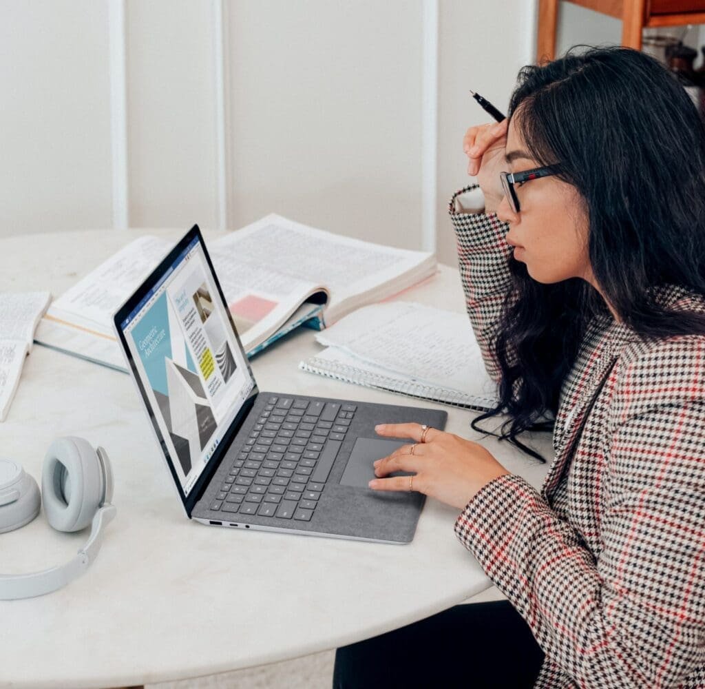 A young woman in glasses, focused on her laptop, reviews architectural designs amid notebooks and study materials on a table.