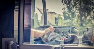 Bus driver's hand reaches for controls inside the cockpit, with buildings and greenery visible through the windshield.