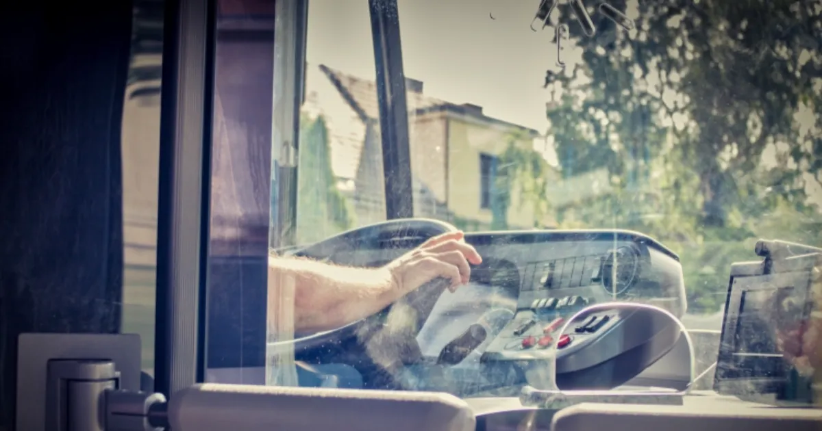 Bus driver's hand reaches for controls inside the cockpit, with buildings and greenery visible through the windshield.