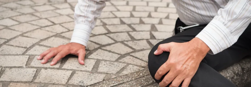 A man in business attire kneels on a stone pavement, holding his knee in pain, suggesting an injury or fall.