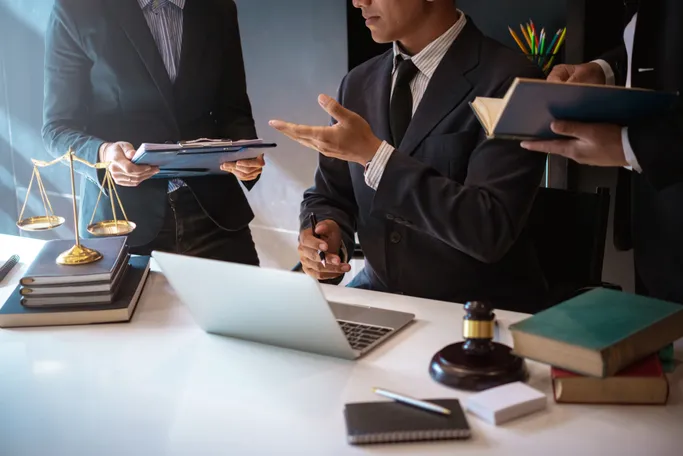 Three lawyers discuss case files and legal documents at a desk with a laptop, books, and a gavel, emphasizing collaboration in legal practice.