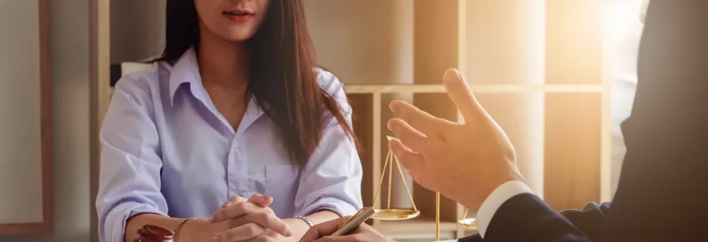 A woman in a light blue shirt is seated at a table, listening intently as a man gestures with his hand. A scale of justice is visible in the background, symbolizing a legal discussion.