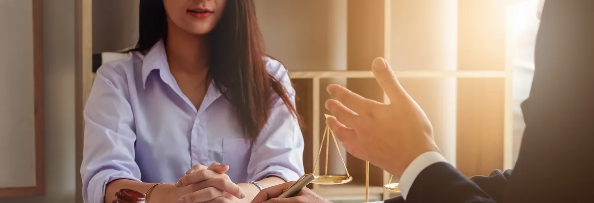 A woman in a light blue shirt is seated at a table, listening intently as a man gestures with his hand. A scale of justice is visible in the background, symbolizing a legal discussion.