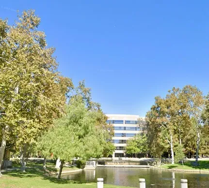 Lush trees frame a serene pond, reflecting a modern office building under a clear blue sky, showcasing a peaceful outdoor workspace.