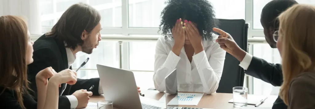 A distressed woman in a business meeting covers her face, while three colleagues discuss around a table with a laptop and documents, indicating conflict or criticism.