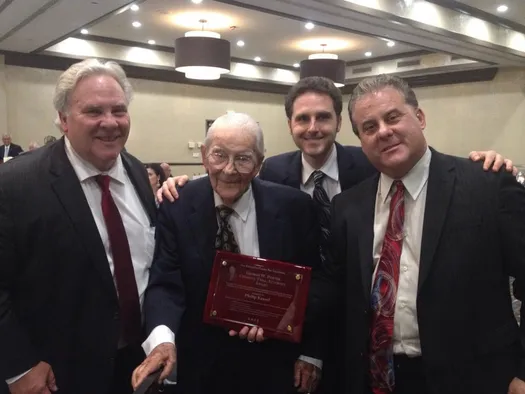 Four men, one elderly, celebrate as he holds an award plaque in a formal event setting, highlighting a moment of recognition and achievement.