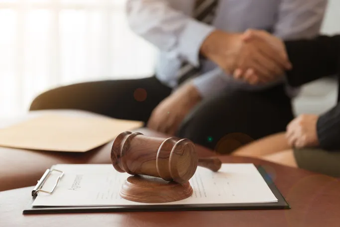 A wooden gavel rests on a legal document as two people shake hands in a law office, symbolizing agreement or contract signing.