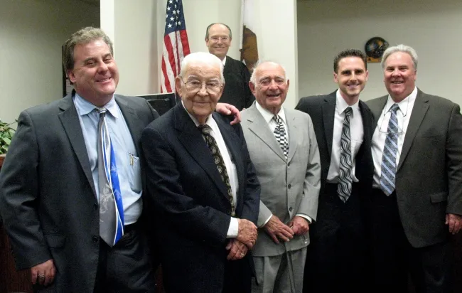 Five men, including an elderly gentleman at center, gather in a formal setting with flags in the background, likely for a special event.