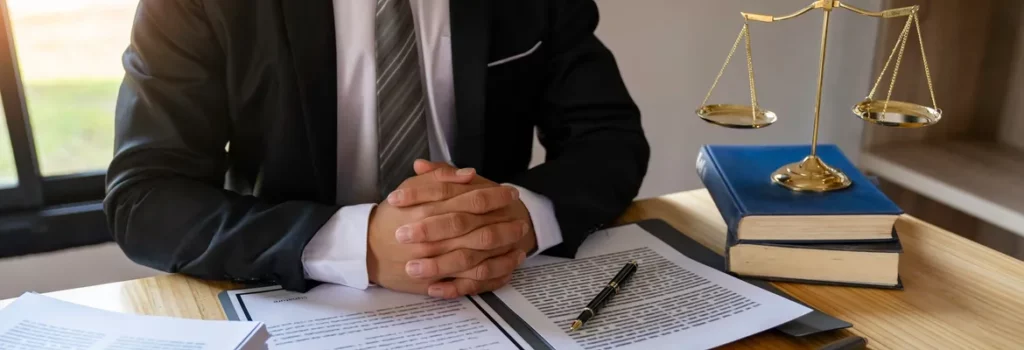 A businessman sits at a desk with clasped hands, surrounded by legal documents, a pen, and a balance scale, reflecting the seriousness of legal matters.