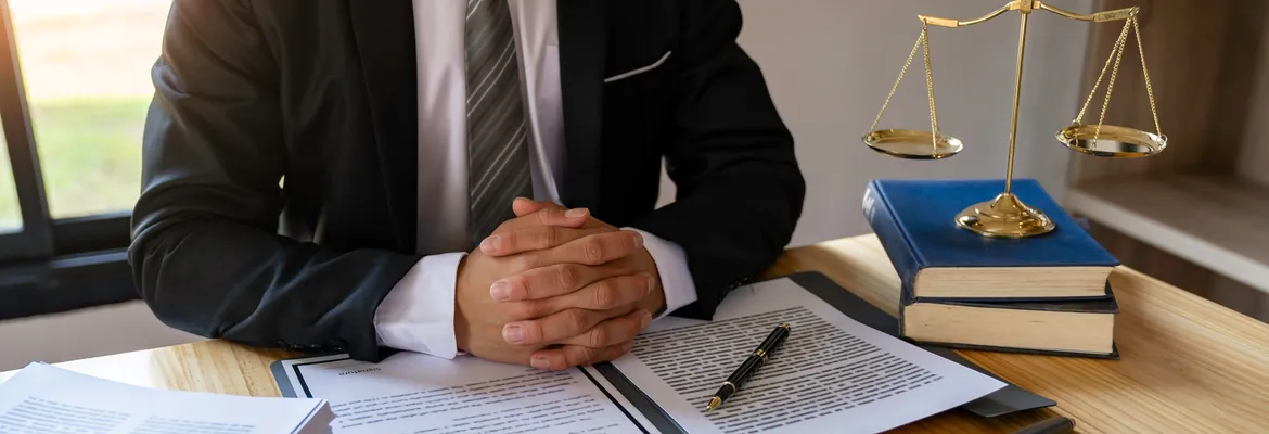 A businessman sits at a desk with clasped hands, surrounded by legal documents, a pen, and a balance scale, reflecting the seriousness of legal matters.