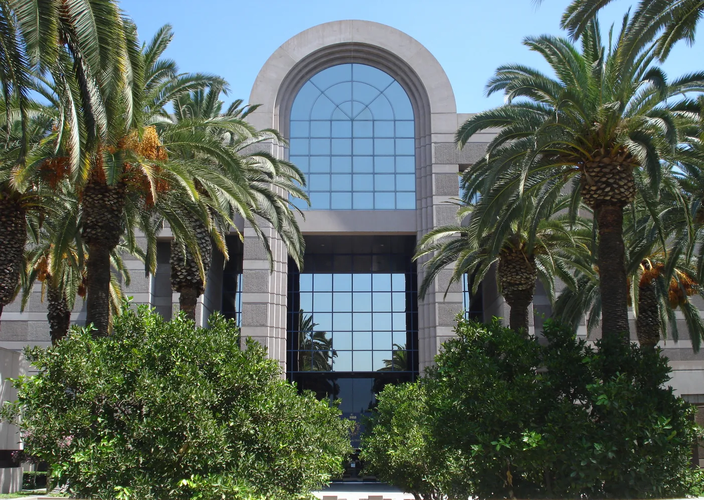 Modern architecture featuring a glass-fronted building framed by palm trees and lush greenery under a clear blue sky.