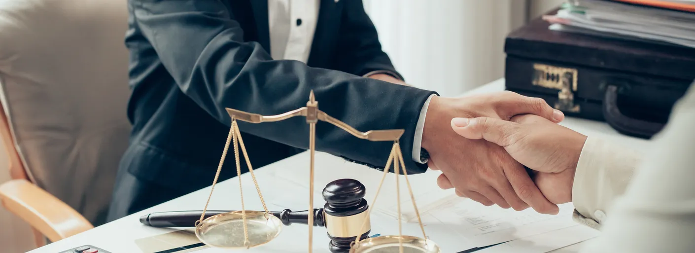 A lawyer and client shake hands over a desk with scales of justice and a gavel, symbolizing agreement and legal partnership.