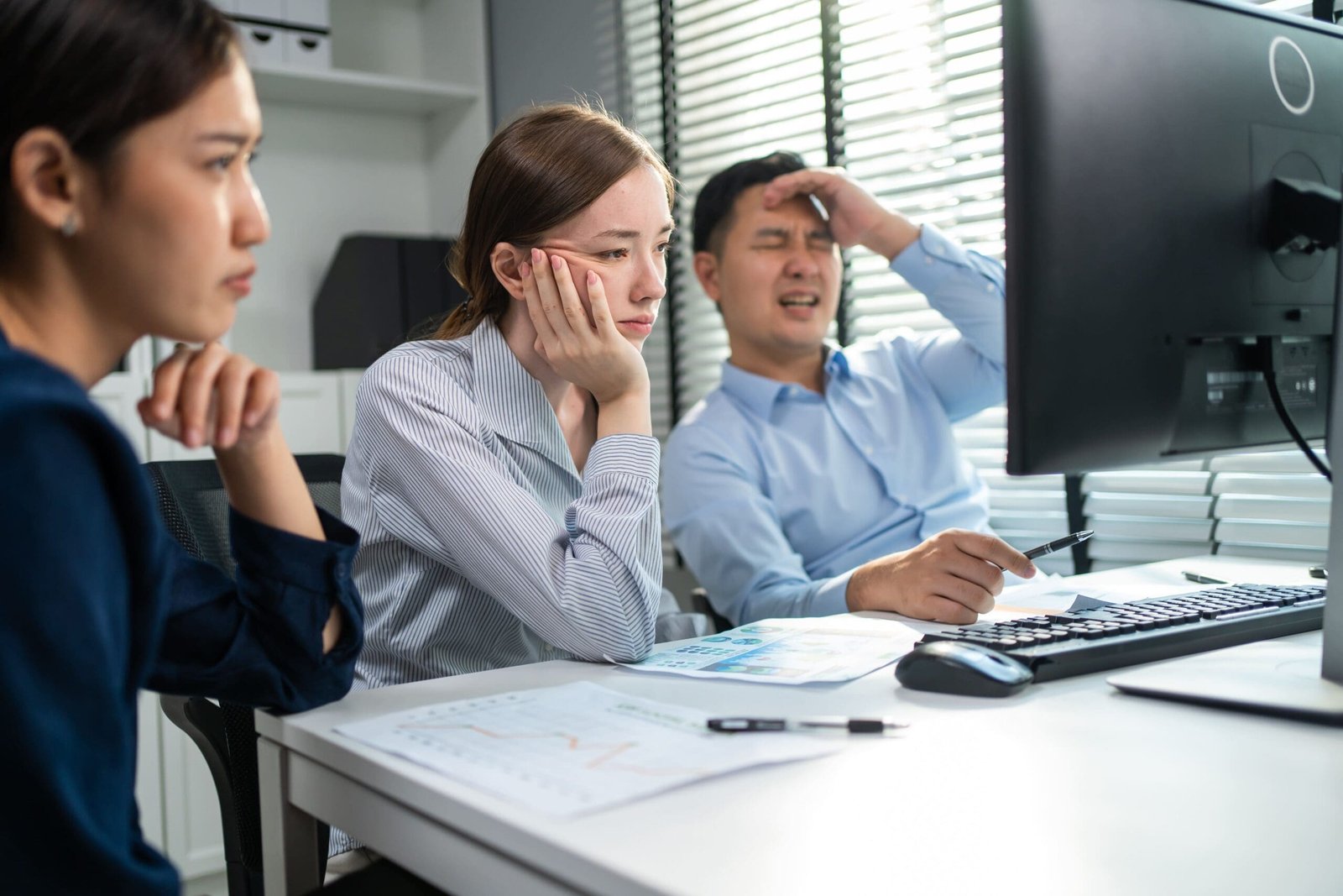 Three colleagues display frustration during a meeting, reviewing financial reports at a desktop computer in a bright office setting.