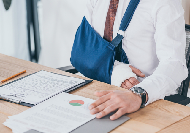 man in white shirt and maroon tie sitting at a desk with his arm in a cast and a blue sling to support it while he tries to be productive at work