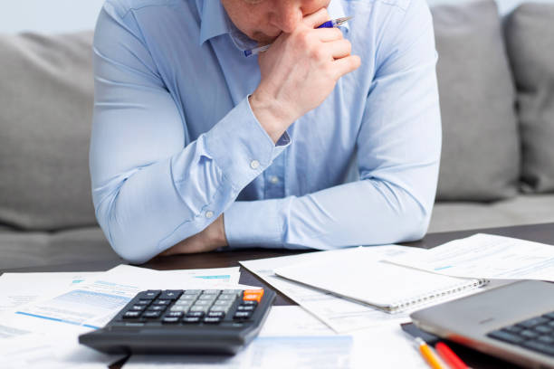 A man in a blue shirt looks stressed while reviewing financial documents and calculations on a calculator, surrounded by paperwork.