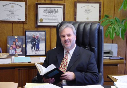 A man in a suit sits in a large leather chair, holding an open book. Behind him are framed diplomas and a family photo, suggesting a scholarly and professional environment.