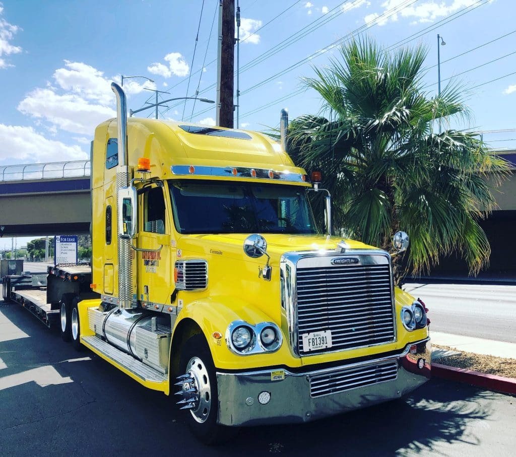 Bright yellow semi-truck parked on a street, showcasing its chrome detailing and large trailer. The truck's vibrant color stands out against a sunny background.