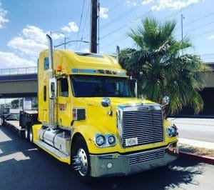 Bright yellow semi-truck parked on a street, showcasing its chrome detailing and large trailer. The truck's vibrant color stands out against a sunny background.